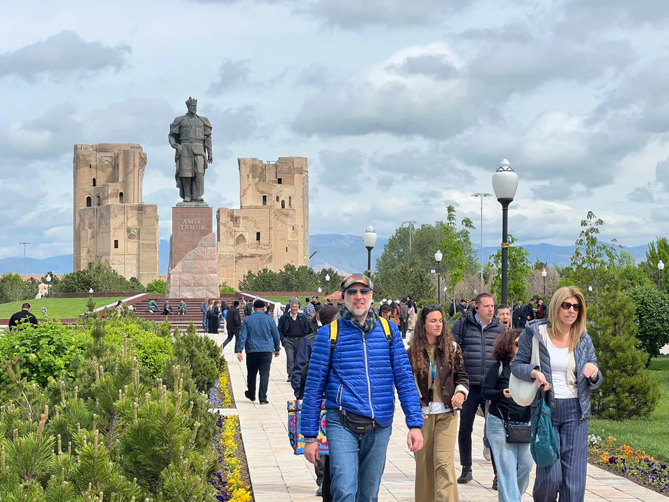 Shahrisabz shahridagi Amir Temur maydonida xorijiy turistlar sayr qilmoqda. Orqa fonda Amir Temur haykali va Oqsaroy saroyi obidasi ko‘rinadi. Qashqadaryo viloyati, Oʻzbekiston.