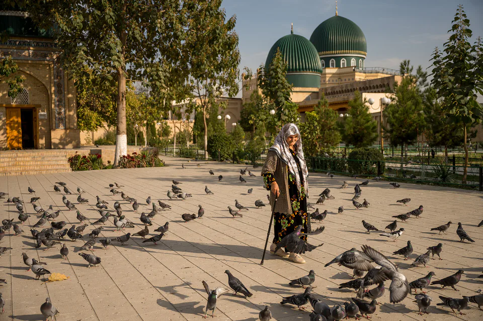 Old woman walks with the cane through the flock of pigeons. Kaptarli mosque on the background. Yurmaduz mahalla, Margilan, Ferghana Region, Uzbekistan.